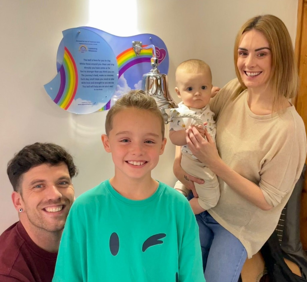 From left to right, dad, son, baby and mum smiling in front of end of cancer treatment bell