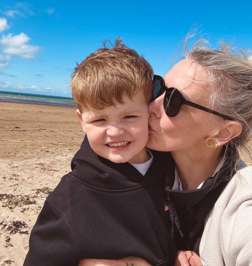 child is smiling at camera with mum on the right who is giving him a kiss on the cheek, they are on the beach on a sunny day